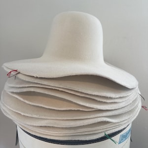 May include: A stack of white felt hats with wide brims and rounded crowns. The hats are stacked on a white and blue cylindrical container. Small red and green threads are visible on the edges of the hats.