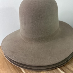 Könnte beinhalten: Ein taupefarbener Hut mit breiter Krempe und hohem, abgerundetem Kopf. Der Hut ist aus einem weichen, filzartigen Material gefertigt. Der Hut liegt auf einer Holzoberfläche. Das Design ist schlicht und klassisch.