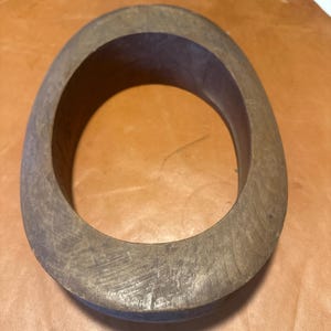 May include: A weathered, oval-shaped wooden bracelet with a dark interior. The bracelet has a rustic, worn appearance, with visible grain and slight imperfections. The color is a muted brown with hints of gray.