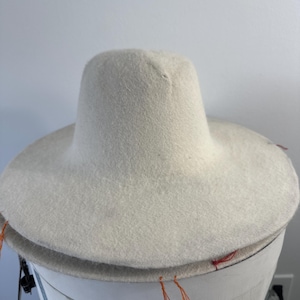 May include: A wide-brimmed, off-white hat with a tall, cylindrical crown. The hat appears to be made of a soft, felt-like material. Orange threads are visible along the brim's edge. The hat is sitting on a white surface.
