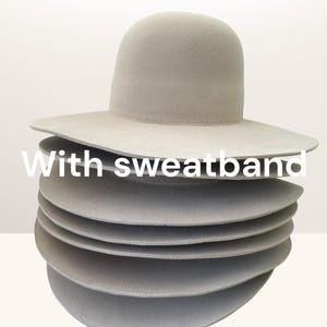 Puede incluir: Una pila de sombreros fedora gris claro con ala ancha. Los sombreros están hechos de un material suave y tienen un diseño clásico. El texto "With sweatband" es visible en la imagen.