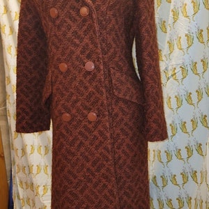 Puede incluir: Un abrigo marrón de doble botonadura con un patrón de chevron. El abrigo tiene cuello y mangas largas.