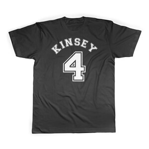 Puede incluir: Camiseta negra con texto blanco que dice "Kinsey 4" en una fuente de estilo universitario.