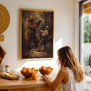 May include: A framed painting of a man interacting with a large brown bear in a forest setting. The painting is hanging on a white wall in a room with a wooden table and fruit on it.