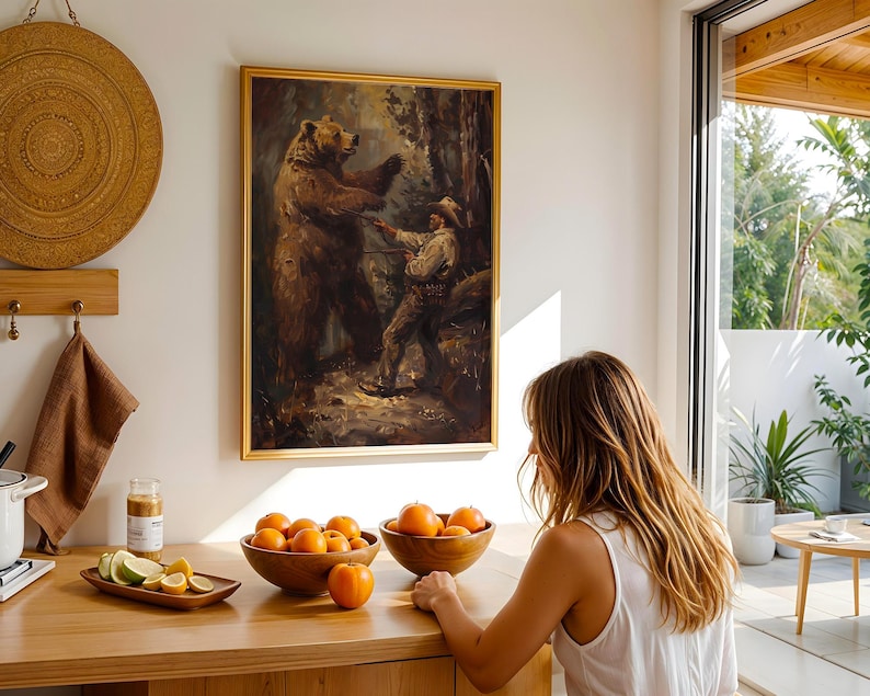 May include: A framed painting of a man interacting with a large brown bear in a forest setting. The painting is hanging on a white wall in a room with a wooden table and fruit on it.