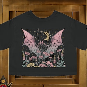May include: Black cropped t-shirt with a pink and gold bat flying over a field of flowers. The bat has a crescent moon above it and stars in the background.