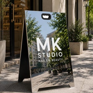 Pode incluir: Uma placa de calçada em forma de A de cor prateada com uma superfície reflexiva apresentando o texto branco MK STUDIO NAILS & BEAUTY. A placa tem uma alça preta recortada no topo e está posicionada em uma calçada pavimentada.