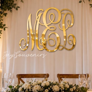 Op de afbeelding: Een groot, goudkleurig, reflecterend monogrambord met de verstrengelde letters M en L in een decoratief scriptlettertype. Het bord is gemonteerd op een witte stoffen achtergrond, met de tekst Jay Souvenirs in het wit onder de letters gedrukt.