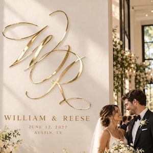 May include: A large gold metallic monogram featuring the letters WR in a cursive script mounted on a white wall. Below the monogram, the text reads WILLIAM & REESE, JUNE 12, 2027, AUSTIN, TX. The display is positioned above a stone table with flowers.
