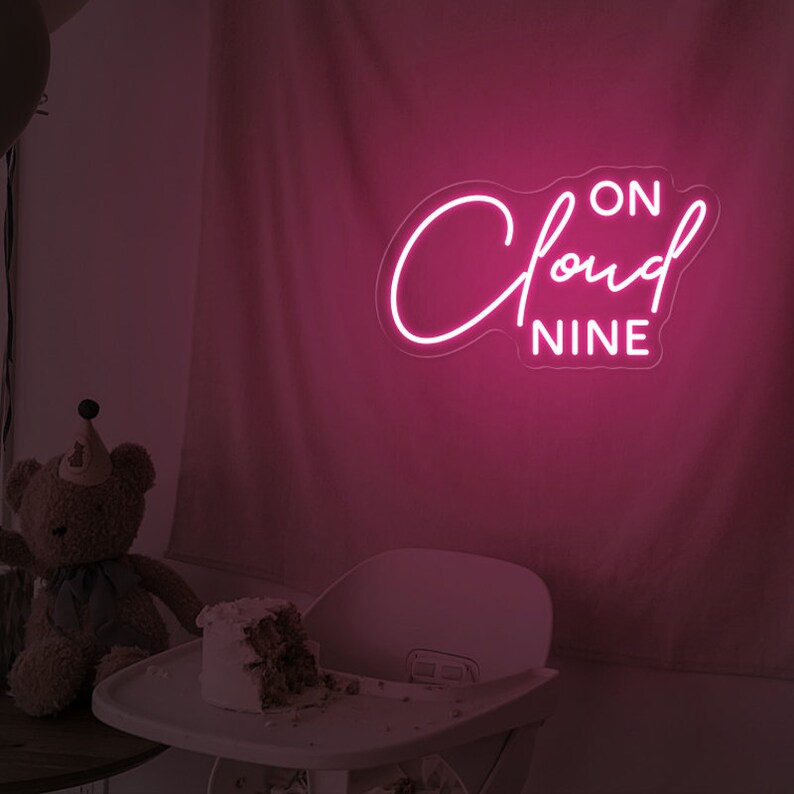 May include: Pink neon sign with the text "ON CLOUD NINE" in a cursive font. The sign is hanging on a pink wall.
