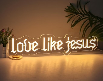 May include: A neon sign with the words "Love Like Jesus" in a stylized font. The sign is made of clear acrylic with warm white LED lights. The sign is on a table with a plant in the background.