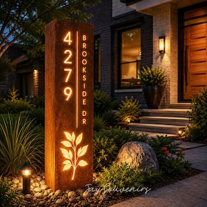 Può includere: Un alto cartello civico rettangolare in metallo color ruggine con intagli illuminati. Il cartello mostra il numero civico verticale 4279 e il testo BROOKSIDE DR, insieme a un motivo a foglia luminoso. La luce brilla dall'interno della struttura.