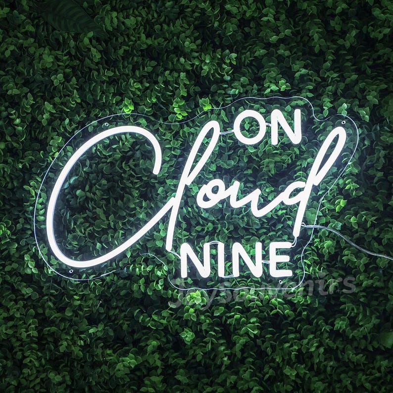 May include: White neon sign with the text "On Cloud Nine" against a green leafy background.