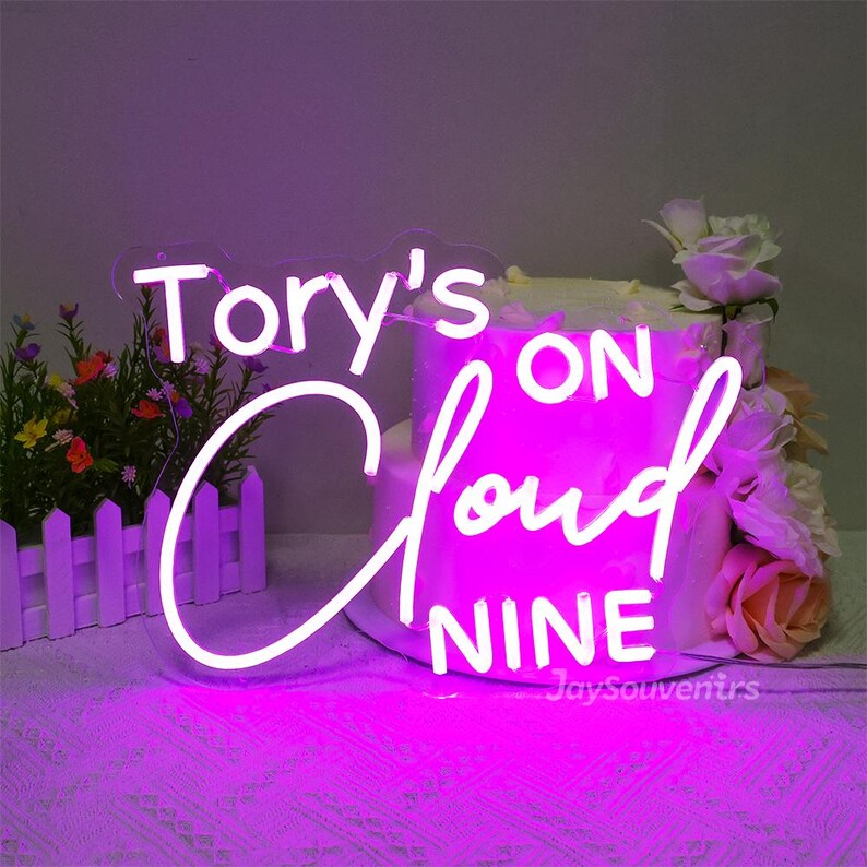 May include: A pink neon sign with the text "Tory's on cloud nine". The sign is lit up and is sitting on top of a white cake.
