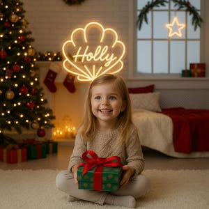 Puede incluir: Una niña sonriente sostiene una caja de regalo verde con una cinta roja. Un letrero de neón sobre su cabeza dice "Holly" en un diseño de concha. Un árbol de Navidad y otras decoraciones festivas están en el fondo.