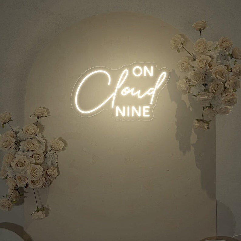 May include: White neon sign with the text "ON CLOUD NINE" on a white background with white flowers on either side.