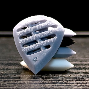 Op de afbeelding: Een stapel gitaar plectrums in verschillende kleuren, waaronder wit, zwart, grijs en een lichtblauwe plectrum met het nummer 4. De lichtblauwe plectrum heeft de tekst "THRPKTHRK" erin gegraveerd. De plectrums zijn gestapeld op een donker houten oppervlak.