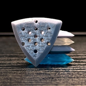 May include: A stack of guitar picks in various colors, including blue, gray, and white. The top pick is a triangular shape with multiple holes and the text "THK PK". The number "4" is visible on the pick.