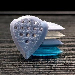 May include: A stack of guitar picks in shades of blue, white, and clear. The top pick is light blue with the text "THIK PK THK" and the number "4". The picks are stacked on a dark, textured surface.