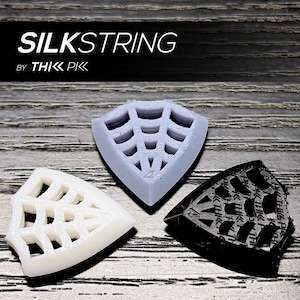 Puede incluir: Tres púas de guitarra en blanco, gris y negro, cada una con un diseño geométrico único. Las púas tienen forma de escudo y presentan un patrón de recortes similar a una telaraña. El texto "SILKSTRING" y "by THIK PIK" se muestran en la parte superior.