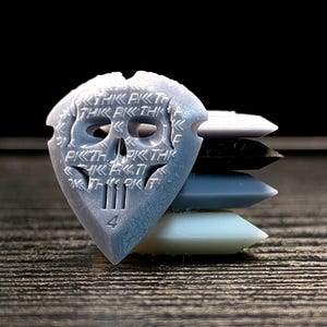 May include: A stack of guitar picks in various colors. The top pick is light gray and features a skull design with the text "PKT" repeated. Below are picks in white, black, light blue, and white. The picks are on a dark wooden surface.