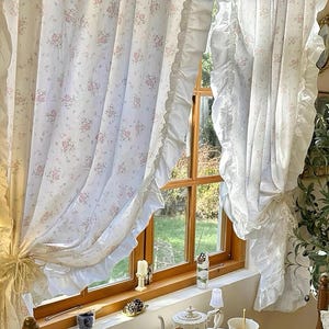 以下が含まれることがあります： ピンクのバラ模様の白い花柄カーテンが窓を縁取っています。カーテンはフリルエッジで、装飾的なリボンで結ばれています。テーブルにはティーポット、ティーカップ、小さなケーキが置かれています。