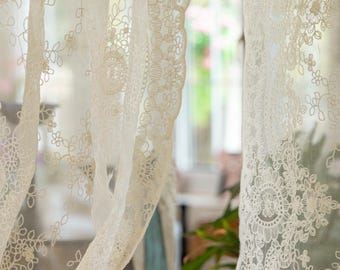 May include: White lace curtains with a floral pattern hang in a window. The curtains are sheer and delicate, and the floral pattern is intricate and detailed.