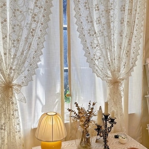 May include: White lace curtains with a floral pattern hang in a window. A yellow lamp with a white shade sits on a table with a white tablecloth. A vase with dried flowers and a candlestick holder are also on the table.