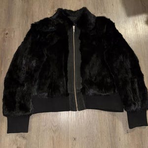 Könnte beinhalten: Eine schwarze Bomberjacke aus Kunstpelz mit Reißverschluss und geripptem Strickkragen, Bündchen und Saum.