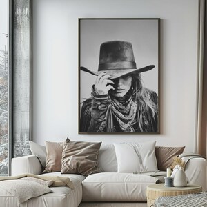 Op de afbeelding: Zwart-wit ingelijste print van een persoon met een cowboyhoed, bandana en jas. De persoon raakt de rand van de hoed aan. De afbeelding hangt boven een witte bank met kussens.