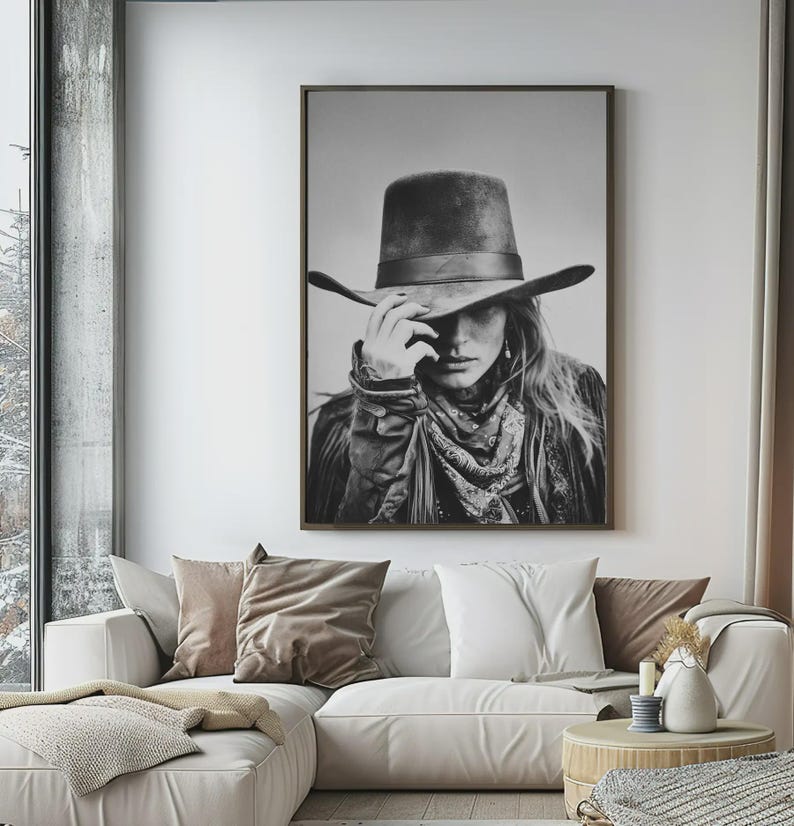 Op de afbeelding: Zwart-wit ingelijste print van een persoon met een cowboyhoed, bandana en jas. De persoon raakt de rand van de hoed aan. De afbeelding hangt boven een witte bank met kussens.