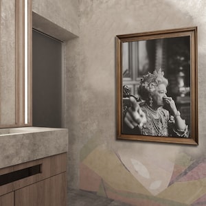 May include: A black and white photograph of a woman wearing a crown and a necklace. She is pointing at the viewer with her right hand and holding her left hand to her nose. The photograph is framed in a brown wooden frame.