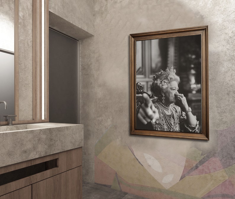 May include: A black and white photograph of a woman wearing a crown and a necklace. She is pointing at the viewer with her right hand and holding her left hand to her nose. The photograph is framed in a brown wooden frame.