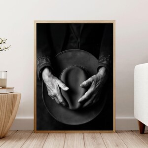 Pu&ograve; includere: Fotografia in bianco e nero di mani che tengono un cappello a tesa larga. Il cappello &egrave; fatto di un materiale scuro e ha una superficie leggermente strutturata. Le mani della persona sono unite intorno al cappello e le sue dita sono visibili.