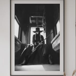May include: Black and white framed print of a person in a train car, wearing a wide-brimmed hat, long coat, and boots, holding a rifle. The image has a vintage, western theme.