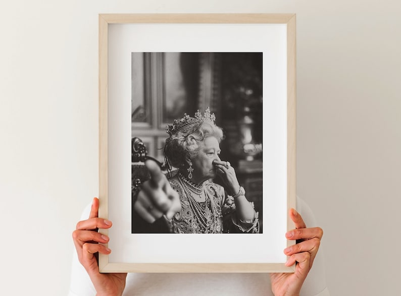 May include: A black and white photograph of a woman wearing a crown and a necklace. She is sitting in a chair and looking at the camera. Her right hand is raised, pointing towards the viewer.
