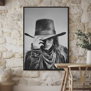 Op de afbeelding: Zwart-wit ingelijste print van een persoon met een cowboyhoed, bandana en jas. De persoon raakt de rand van de hoed aan. De afbeelding wordt weergegeven op een stenen muur.