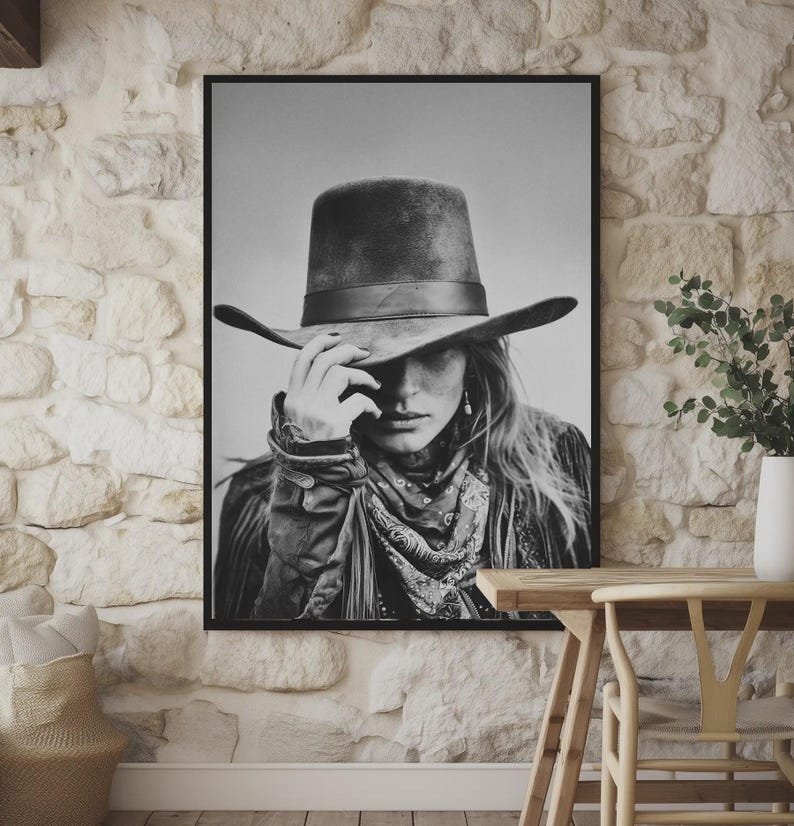 Op de afbeelding: Zwart-wit ingelijste print van een persoon met een cowboyhoed, bandana en jas. De persoon raakt de rand van de hoed aan. De afbeelding wordt weergegeven op een stenen muur.