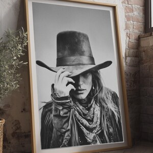 Op de afbeelding: Zwart-wit ingelijste print van een persoon met een cowboyhoed, bandana en jas. De afbeelding leunt tegen een bakstenen muur en een houten vloer. De persoon raakt de rand van de hoed aan.