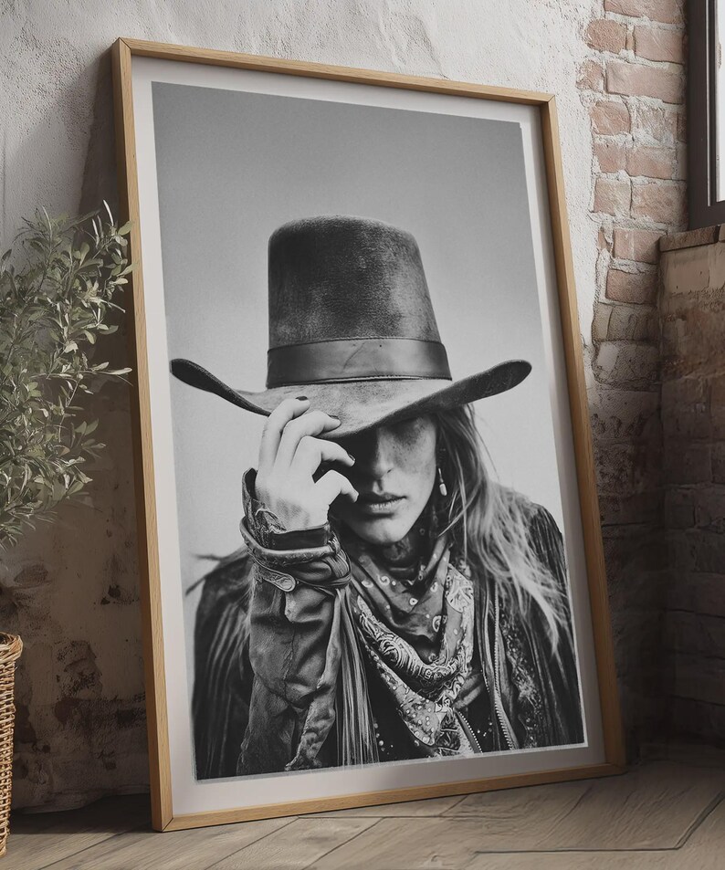 Op de afbeelding: Zwart-wit ingelijste print van een persoon met een cowboyhoed, bandana en jas. De afbeelding leunt tegen een bakstenen muur en een houten vloer. De persoon raakt de rand van de hoed aan.