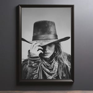 Op de afbeelding: Zwart-wit ingelijste print van een persoon met een brede hoed, een patroon sjaal en een jas. Het gezicht is gedeeltelijk bedekt door de hoed, met een hand die de rand aanraakt. De afbeelding heeft een vintage, rustieke uitstraling.