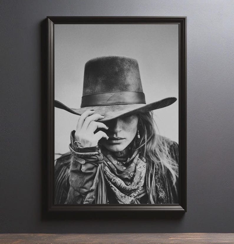 Op de afbeelding: Zwart-wit ingelijste print van een persoon met een brede hoed, een patroon sjaal en een jas. Het gezicht is gedeeltelijk bedekt door de hoed, met een hand die de rand aanraakt. De afbeelding heeft een vintage, rustieke uitstraling.