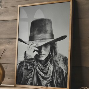 Op de afbeelding: Zwart-wit ingelijste print van een persoon met een breedgerande hoed, een patroon sjaal en een jas. De persoon raakt de rand van de hoed aan. De afbeelding heeft een vintage, rustieke uitstraling.