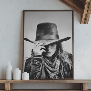 Op de afbeelding: Zwart-wit ingelijste print van een persoon met een brede cowboyhoed, met hun hand op de rand. De persoon draagt ook een sjaal met patroon en een jas. De print wordt tentoongesteld op een houten plank.