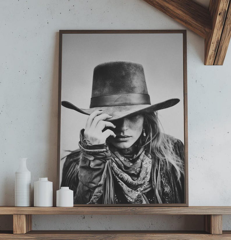 Op de afbeelding: Zwart-wit ingelijste print van een persoon met een brede cowboyhoed, met hun hand op de rand. De persoon draagt ook een sjaal met patroon en een jas. De print wordt tentoongesteld op een houten plank.