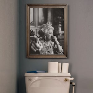 May include: A black and white framed photo of a woman wearing a crown and a pearl necklace. She is holding her nose with her right hand and pointing with her left hand.