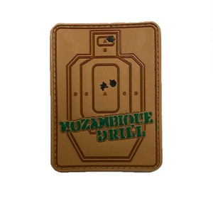 May include: A tan-colored patch with a target silhouette and the text "MOZAMBIQUE DRILL" in green. The target has two black bullet holes.