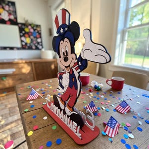 Puede incluir: Una figura de madera de Mickey Mouse con atuendo patri&oacute;tico, con una combinaci&oacute;n de colores rojo, blanco y azul. La figura est&aacute; sobre una base roja con el nombre "EMMANUEL" en blanco. Peque&ntilde;as banderas americanas y confeti.