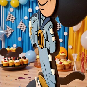 May include: A layered wooden Mickey Mouse figure in a blue and gold outfit, with a gold medal that has the number 1 on it. The figure is part of a party decoration, with cupcakes and balloons in the background.