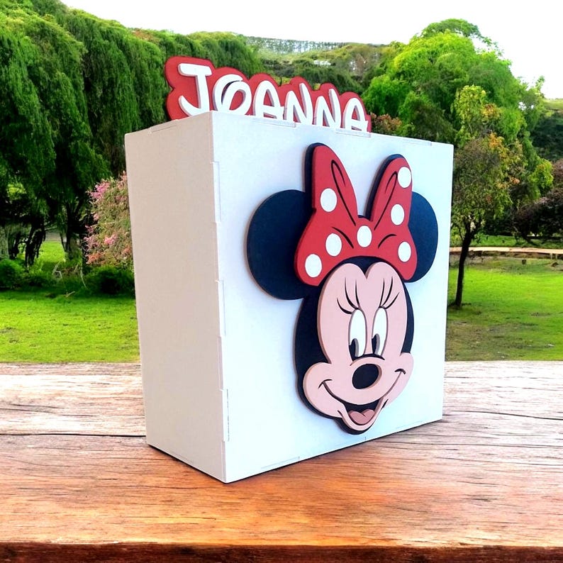 Op de afbeelding: Een witte doos met een Minnie Mouse-ontwerp, inclusief een rode strik met witte stippen. De naam "Joanna" staat in het rood bovenaan. De doos staat op een houten oppervlak met een groene achtergrond.